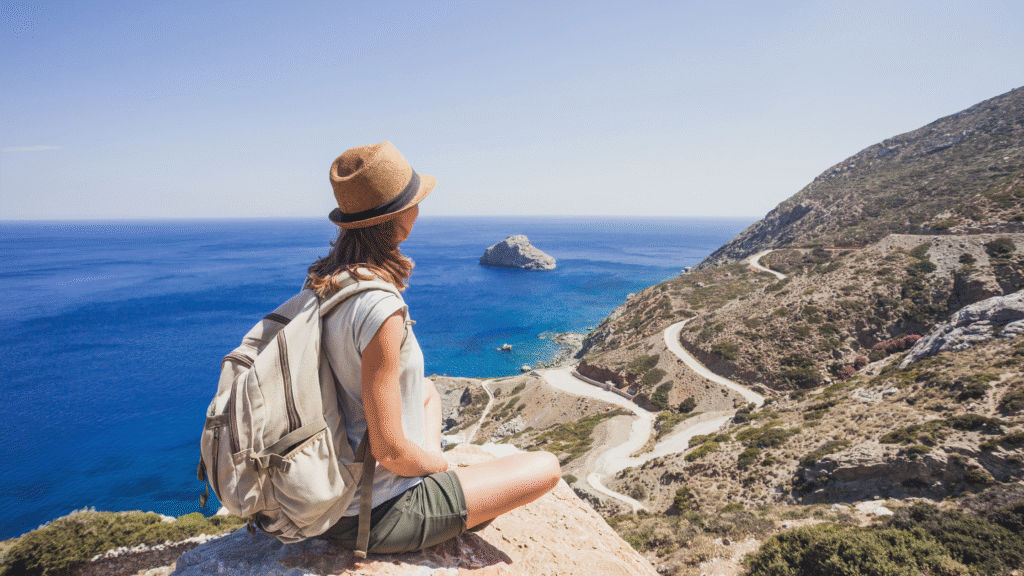 Woman sitting on rocky coastline overlooking the sea, planning eco travel using AI and VR tools, 2025